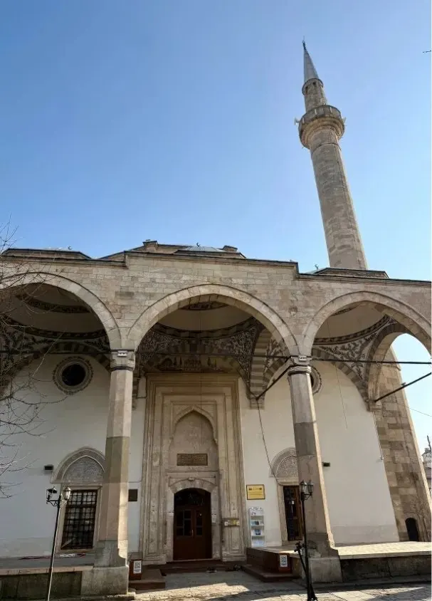 Figure 9 · exterior of an Ottoman-era mosque in Pristina, Kosovo, its arched portico and single minaret against a clear sky