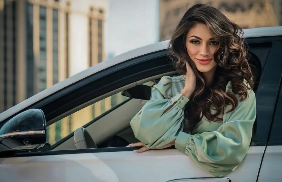 Jillian Batchelor leaning from the window of her Mercedes-Benz S 580 in a soft green satin blouse, hair in voluminous curls, city towers behind