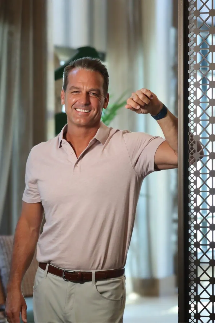 Andrew Jarosz in a pale blush polo shirt, stone trousers and a brown leather belt, leaning against a carved openwork screen in daylight