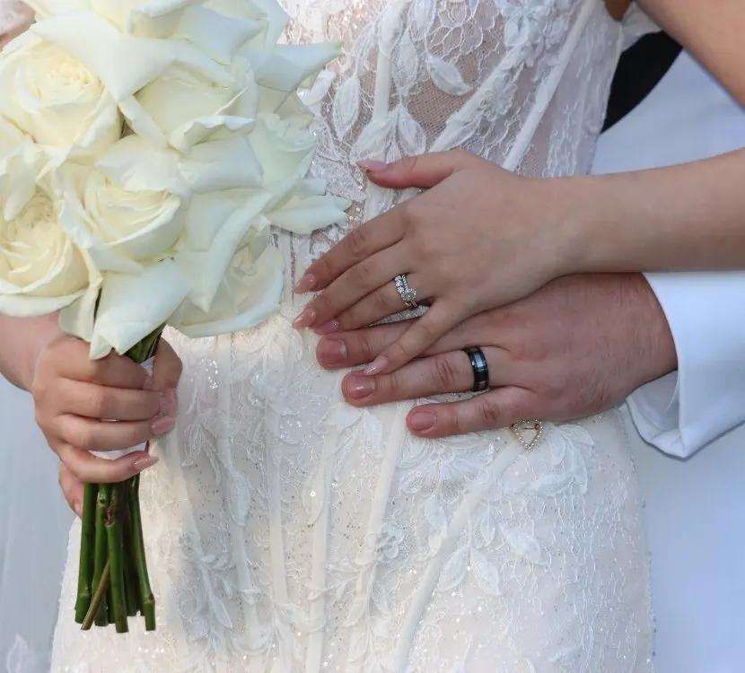 Detail of a bride's and groom's hands at her waist · her pavé diamond ring and his black band against the lace of her gown, a white rose bouquet at the frame's edge
