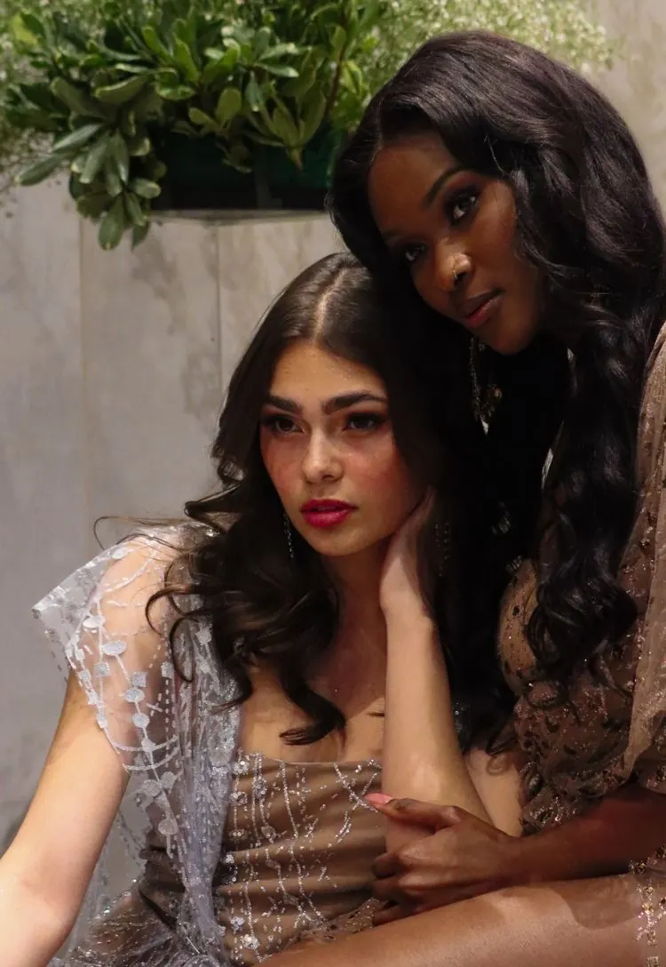 Two models in embellished evening gowns, an editorial portrait: one seated in a silver-beaded gown with a sheer lace overlay, the other standing behind her in a bronze beaded gown, long dark waves, greenery above them