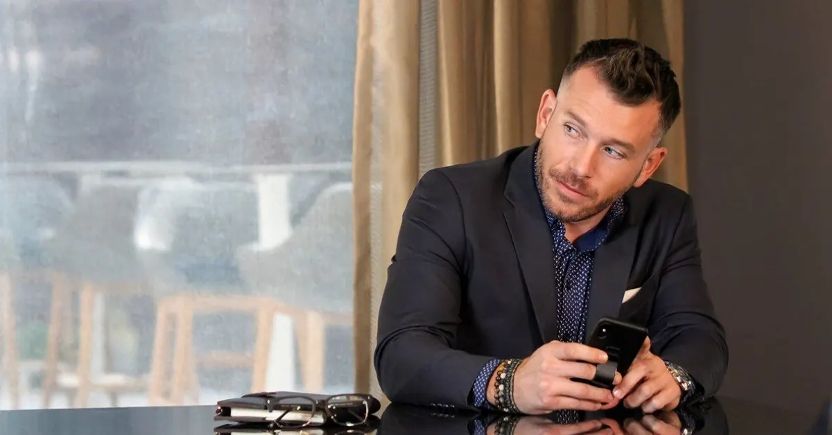 Chris Najarro in a deep navy suit over a dark shirt patterned in small dots, a phone in his hands, beaded bracelets and a watch at his wrist