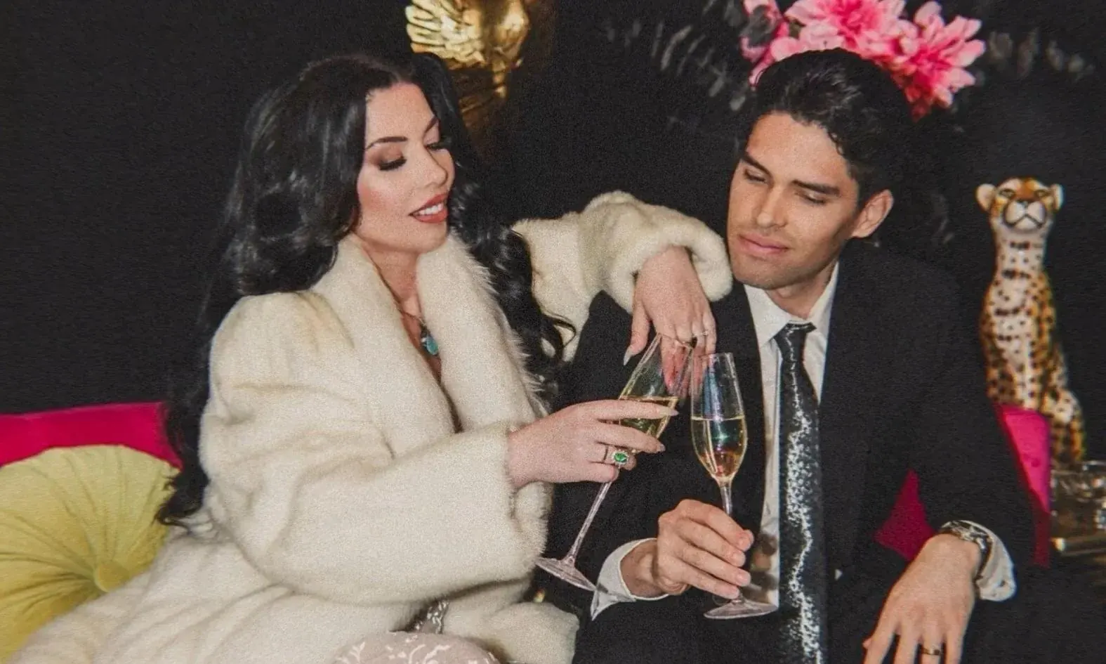Royilda Medina in a white fur coat and Marlon A. Medina in a black suit, seated together and raising champagne glasses in a toast