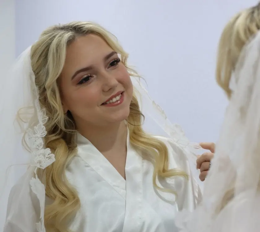 The bride in a satin robe and lace-edged veil, smiling at her reflection in the bridal suite
