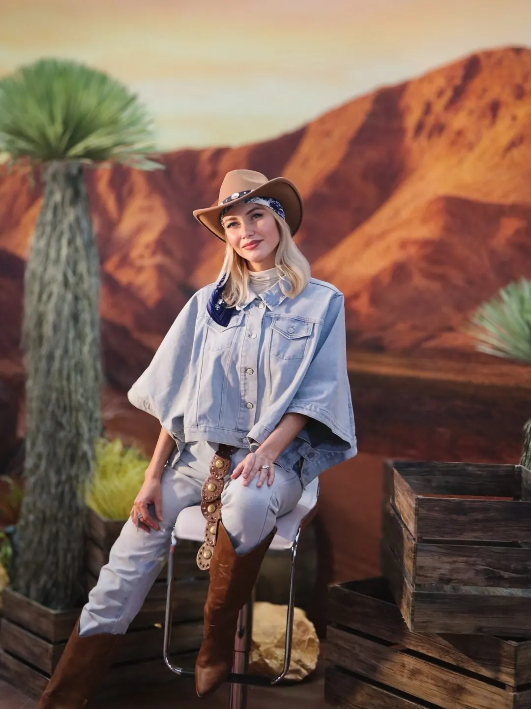 A model in a full western editorial look: washed-denim cape jacket, cream turtleneck, pale denim jeans, concho belt, tan hat and brown knee-high boots, seated in a staged desert set of built red-rock scenery