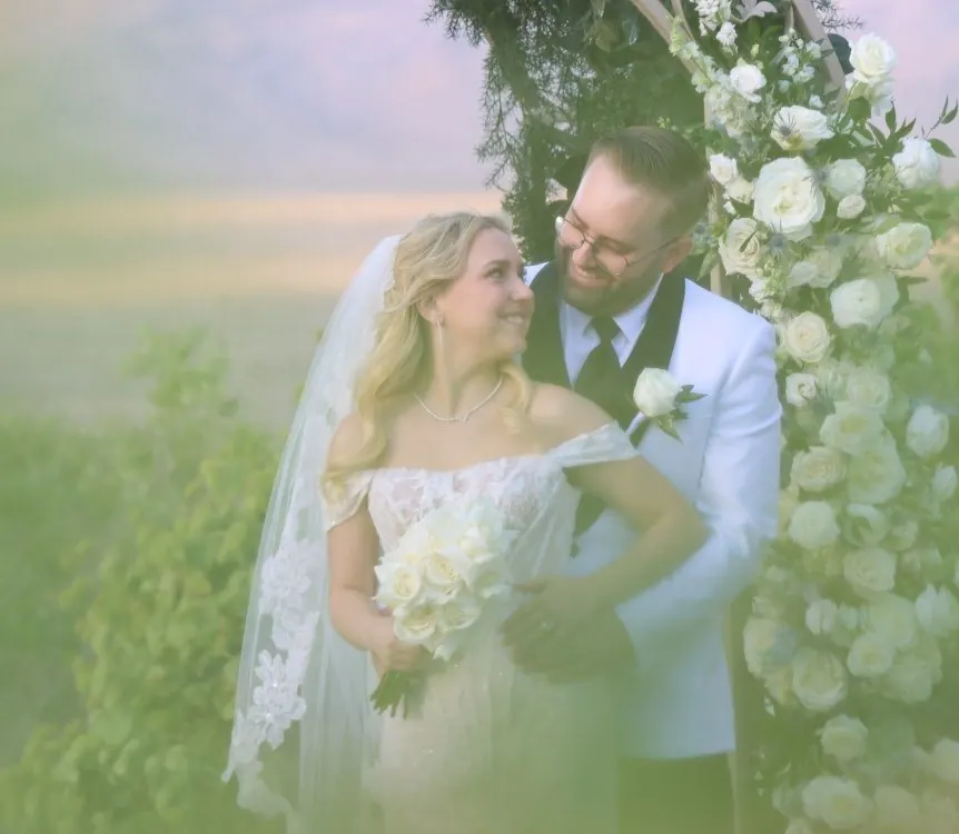 Bride and groom embracing beneath a floral arch of white roses, desert and mountains soft behind them at Paiute Golf Resort